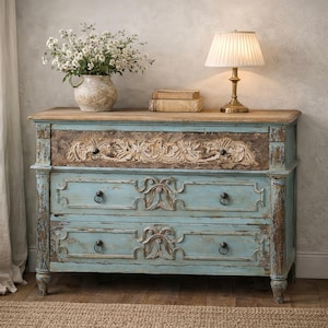 May include: A distressed, light blue chest of drawers with two drawers and a carved floral design. The top is a light wood colour. A lamp with a pleated shade and a vase of white flowers sit on top. The chest has a rustic, antique appearance.