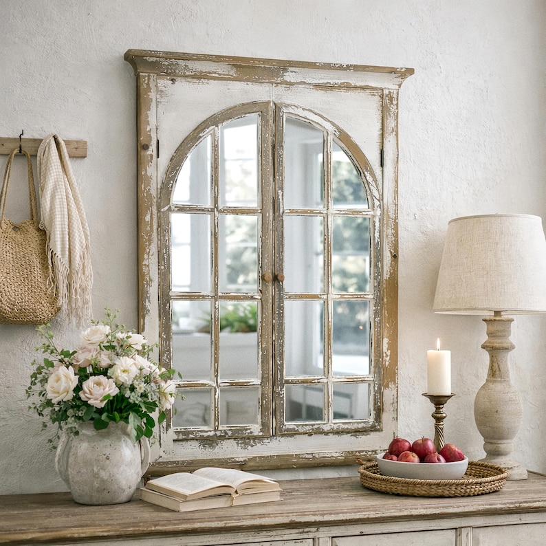 May include: A distressed white and brown arched window mirror with multiple panes. The mirror is mounted on a wall above a wooden table. A vase of flowers, a book, and a lamp are on the table.