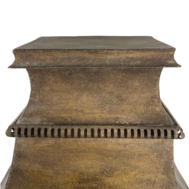 May include: Close-up of a weathered, bronze-colored metal lantern. The lantern has a rectangular top and a decorative base with a series of small, evenly spaced rectangular cutouts. The metal has a textured, aged appearance.