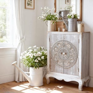 May include: A white distressed cabinet with a decorative circular design on the doors. A white watering can and various metal containers hold white flowers. The scene is set in a bright, sunlit room with a wooden floor.