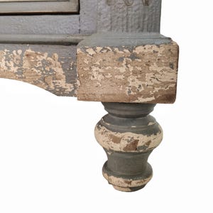 May include: Close-up of a distressed, antique furniture leg. The leg features a turned design with a gray-blue paint finish, showing signs of wear and chipping, revealing the natural wood underneath. The base is a bulbous shape.