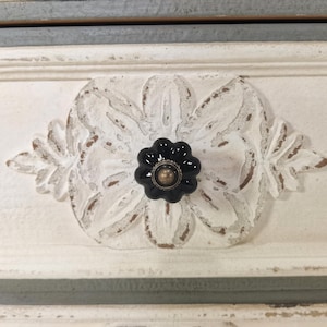 May include: Close-up of a white painted drawer front with a carved floral design and a black flower-shaped knob with a gold center. The drawer has a distressed, vintage look with visible wood grain.