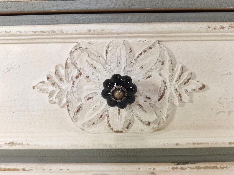 May include: Close-up of a white painted drawer front with a carved floral design and a black flower-shaped knob with a gold center. The drawer has a distressed, vintage look with visible wood grain.