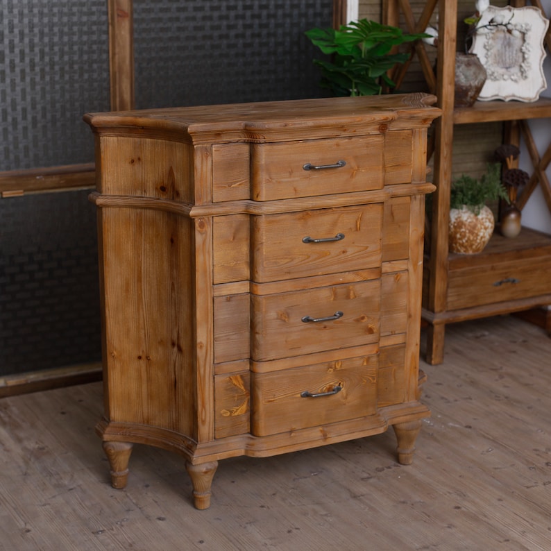May include: A wooden chest of drawers with five drawers and ornate detailing. The drawers have black metal handles. The chest is a light brown colour and has a distressed finish.