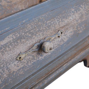 Op de afbeelding: Close-up van een verweerde blauw en bruin houten lade. De lade heeft een decoratieve metalen handgreep met een ronde knop. De verf is afgebladderd, wat het een rustieke, vintage uitstraling geeft. De lade maakt deel uit van een groter meubelstuk.