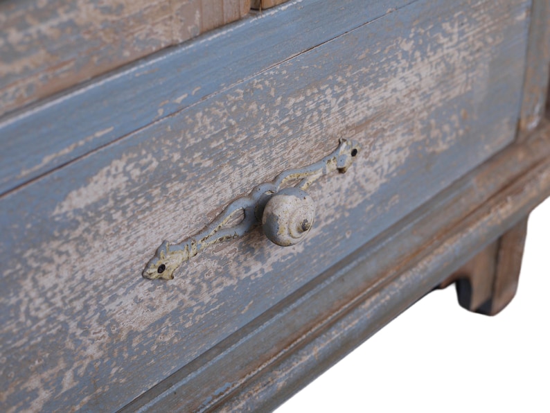 Op de afbeelding: Close-up van een verweerde blauw en bruin houten lade. De lade heeft een decoratieve metalen handgreep met een ronde knop. De verf is afgebladderd, wat het een rustieke, vintage uitstraling geeft. De lade maakt deel uit van een groter meubelstuk.