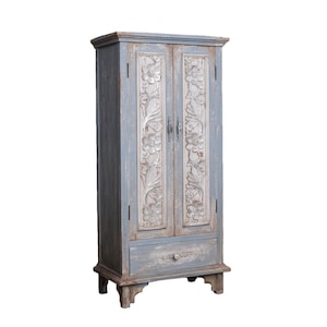 Op de afbeelding: Een hoge, blauw geverfde houten kast met gesneden bloemmotieven op de deuren. De kast heeft een enkele lade aan de onderkant en staat op vier korte poten. De afwerking van de kast heeft een verweerde uitstraling.