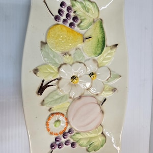 May include: Rectangular ceramic serving platter with a raised relief design of fruit and flowers. The design includes a yellow pear, green pear, purple grapes, white flowers, and a pink peach. The platter has a cream-colored background.