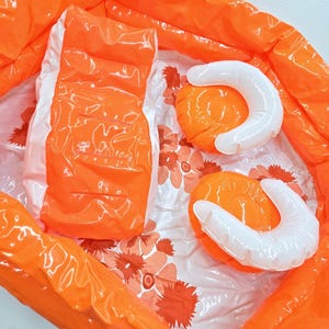 May include: An orange inflatable bag contains an orange rectangular inflatable object and two orange and white inflatable objects. The objects are on a white surface with orange floral patterns. The bag is made of a transparent material.