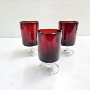 May include: Three ruby red wine glasses with clear stems and bases. The glasses are cylindrical with a slightly flared rim. The glasses are arranged in a triangular pattern against a white background.