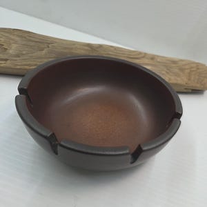 May include: A brown ashtray with a dark rim and three notches for resting cigarettes. The ashtray has a smooth, glossy finish and a circular shape. A piece of driftwood is in the background.