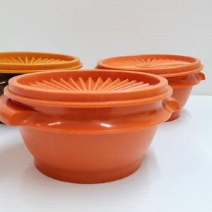 May include: Three orange and brown plastic containers with lids. The containers are round and have a slight curve to them. The lids have a raised design.