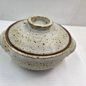 May include: A round ceramic lidded bowl featuring a speckled off-white glaze with brown flecks throughout. The bowl has two small side handles and a matching lid with a central knob, both accented by a dark brown rim along the edges.