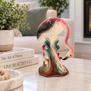 May include: A ceramic flamingo figurine featuring a glossy finish with shades of pink, black, and white. The bird is depicted in a downward-facing pose, perched atop a stylised base coloured in deep green and brown tones with textured details.