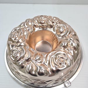 May include: A silver-colored bundt cake pan with a rose design. The pan has a circular shape with a central hole and a decorative floral pattern around the edges. The pan sits on a silver-colored base.