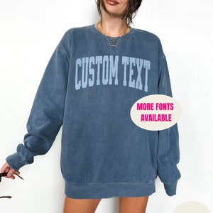 May include: A blue sweatshirt with the text "CUSTOM TEXT" in a light blue color. The sweatshirt has a crew neck and long sleeves. The text "MORE FONTS AVAILABLE" is in a white circle on the bottom right of the sweatshirt. The sweatshirt is on a white background.