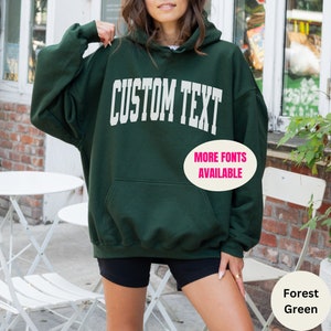 May include: Forest green hooded sweatshirt with the text "CUSTOM TEXT" in white. The text "MORE FONTS AVAILABLE" is also in white.