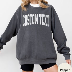May include: A charcoal gray sweatshirt with the text "CUSTOM TEXT" printed in white on the front. The sweatshirt has a relaxed fit and a crew neck.