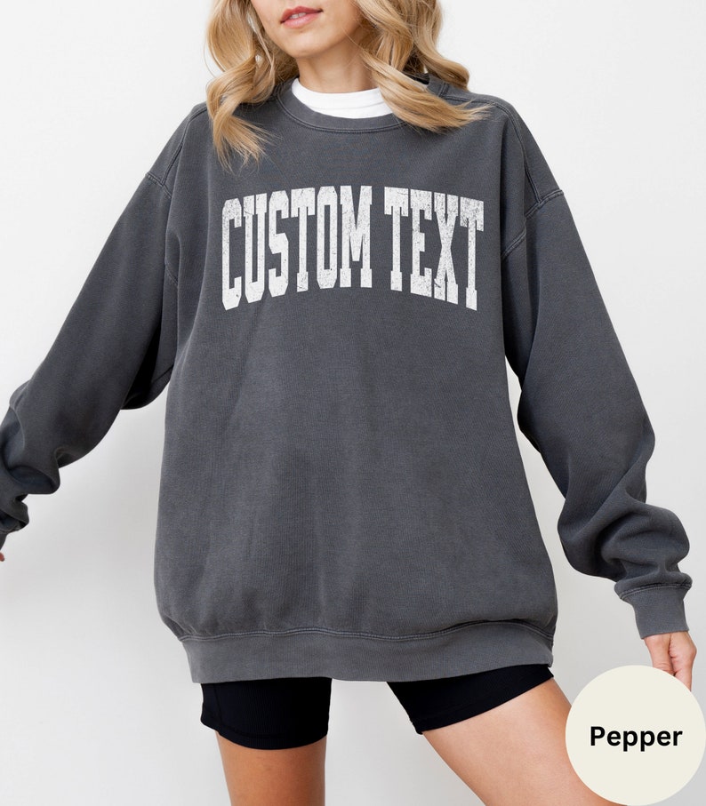 May include: A charcoal gray sweatshirt with the text "CUSTOM TEXT" printed in white on the front. The sweatshirt has a relaxed fit and a crew neck.
