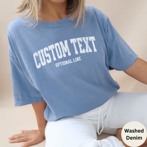 May include: A light blue t-shirt with the text "CUSTOM TEXT" in white, with the words "OPTIONAL LINE" underneath. The t-shirt is made of a soft, washed denim fabric.