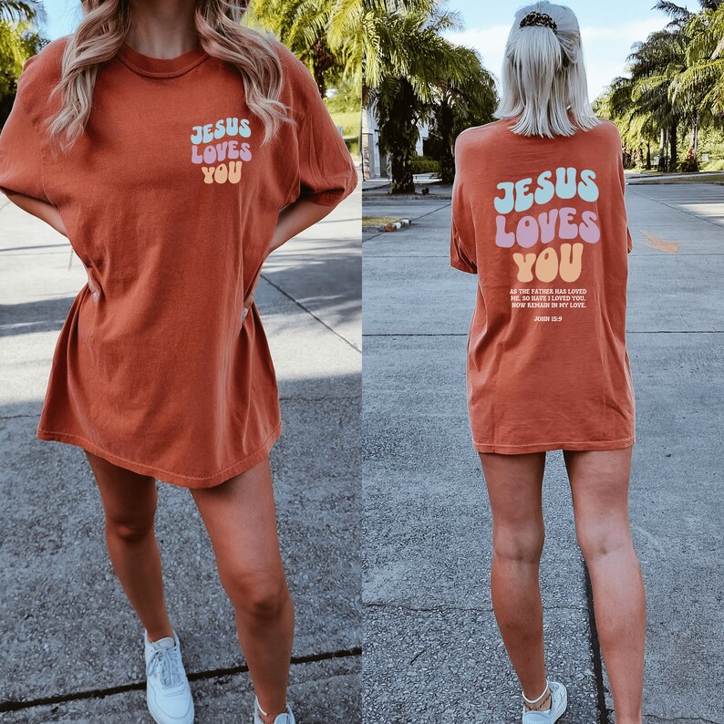 May include: A rust-colored oversized t-shirt with the text "Jesus Loves You" in rainbow-colored letters. The text is in a retro font style. The back of the shirt has the same text with a bible verse from John 15:9.