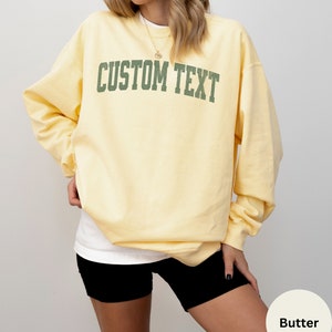 May include: A yellow sweatshirt with the text "CUSTOM TEXT" in green letters. The sweatshirt is being worn by a person.
