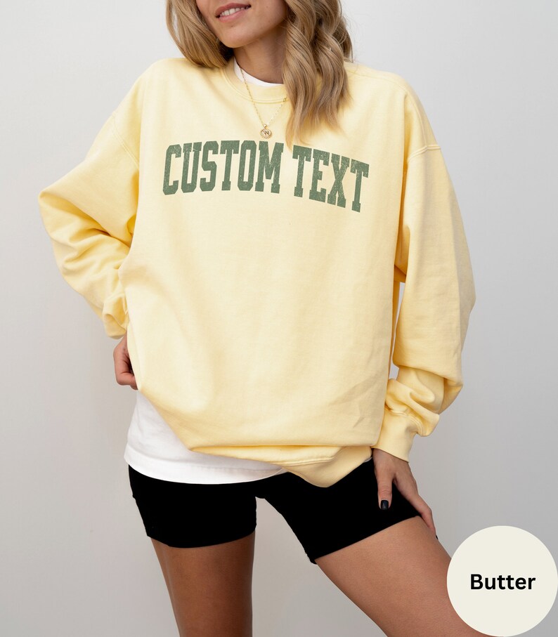 May include: A yellow sweatshirt with the text "CUSTOM TEXT" in green letters. The sweatshirt is being worn by a person.