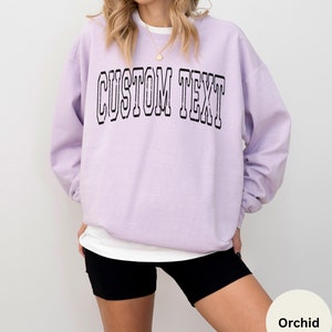 May include: A lavender sweatshirt with the text "CUSTOM TEXT" in black outlined letters.