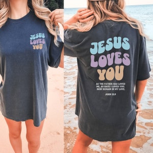 May include: A dark blue t-shirt with the text "Jesus Loves You" in colorful, bubble-like letters. The back of the shirt has the same text with a Bible verse from John 15:9.