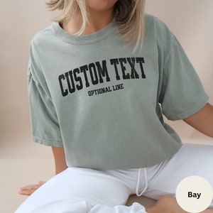 May include: A sage green t-shirt with a black graphic that says "CUSTOM TEXT" and "OPTIONAL LINE" in a vintage collegiate style font.