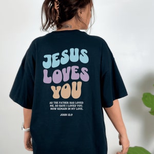 May include: Black t-shirt with a colorful graphic that says "JESUS LOVES YOU" in a retro font. The text "AS THE FATHER HAS LOVED ME, SO HAVE I LOVED YOU. NOW REMAIN IN MY LOVE. JOHN 15:9" is printed below the graphic.