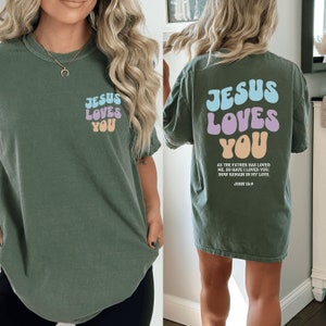 May include: A green t-shirt with the text "Jesus Loves You" in colorful bubble letters on the front and back. The back also includes the scripture John 15:9.