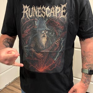Pode incluir: Camiseta preta com uma ilustração de fantasia. O design inclui a palavra "RUNESCAPE" em uma fonte gótica, um mago tocando guitarra, dragões e raios. A camiseta é feita de um material macio.