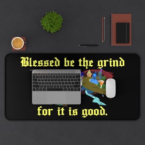 May include: Black desk mat with the text "Blessed be the grind for it is good." in yellow. The mat features a cartoon illustration of a group of people in a pile of blankets.