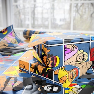 Könnte beinhalten: Zwei Geschenkboxen, verpackt in blauem Papier im Comic-Stil mit farbenfrohen Illustrationen eines Wrestlers. Die Boxen sind auf einer passenden Geschenkpapieroberfläche gestapelt, mit einer Schere und einem schwarzen Band in der Nähe.