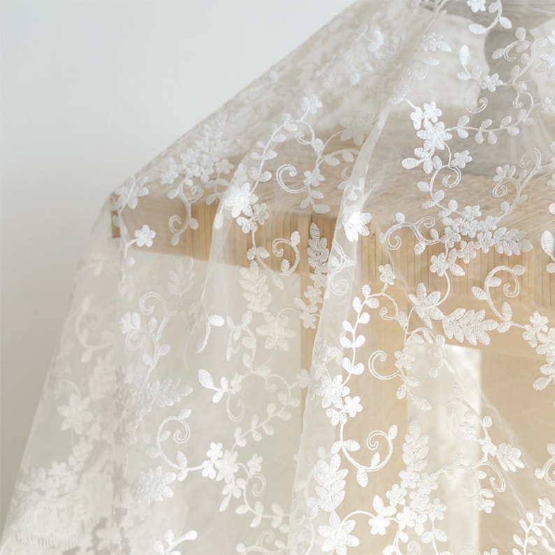 May include: White sheer fabric with a delicate floral embroidery pattern. The fabric is draped over a wooden surface.