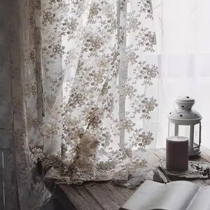 Op de afbeelding: Witte kantgordijnen met een bloemenpatroon hangen in een raam. Een boek en een kaars staan op een houten tafel voor het raam.