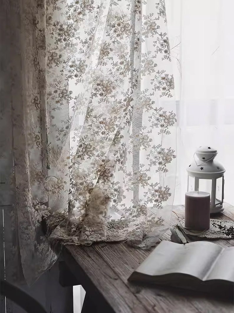Op de afbeelding: Witte kantgordijnen met een bloemenpatroon hangen in een raam. Een boek en een kaars staan op een houten tafel voor het raam.