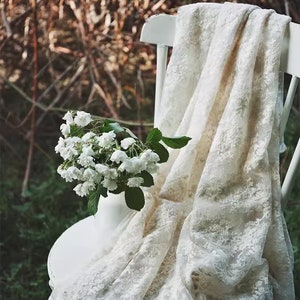 Op de afbeelding: Een witte kanten stof gedrapeerd over een witte stoel met een boeket witte bloemen in een witte vaas.