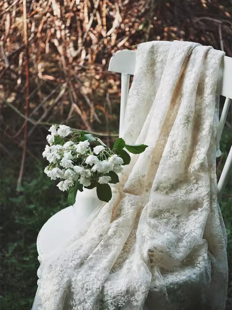 Op de afbeelding: Een witte kanten stof gedrapeerd over een witte stoel met een boeket witte bloemen in een witte vaas.