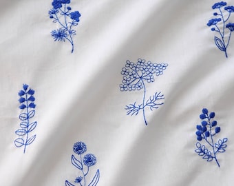 May include: White fabric with a blue embroidered floral pattern. The flowers are stylised and include small clusters of flowers and larger, more open blooms.