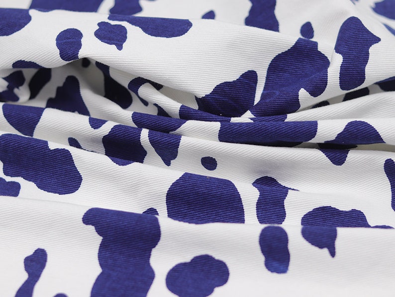 May include: White fabric with a blue cow print pattern. The fabric is folded and wrinkled.