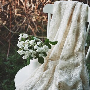 Op de afbeelding: Een witte kanten stof gedrapeerd over een witte stoel met een boeket witte bloemen in een vaas op de zitting.