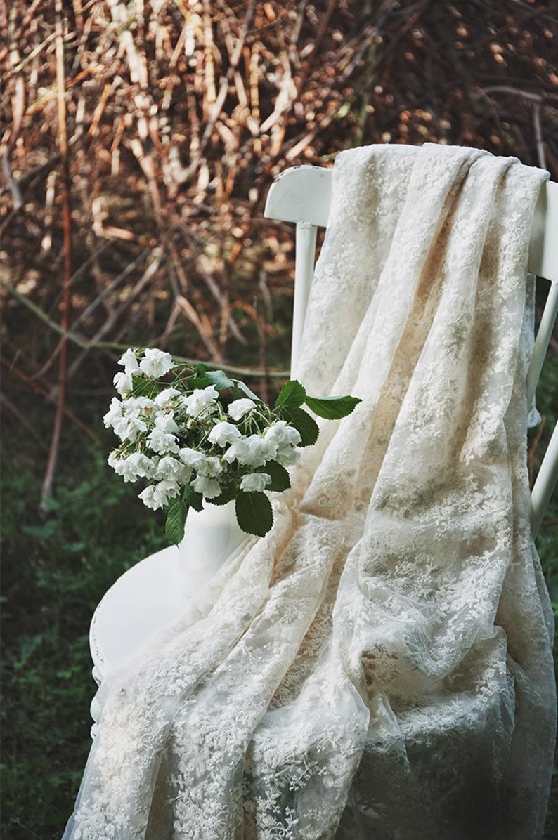 Op de afbeelding: Een witte kanten stof gedrapeerd over een witte stoel met een boeket witte bloemen in een vaas op de zitting.