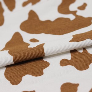 May include: White fabric with a brown cow print pattern. The fabric is folded in half, showing the texture of the material.