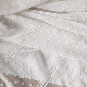 May include: White cotton fabric with a delicate embroidered pattern of small dots and floral designs. The fabric has a slightly textured surface.