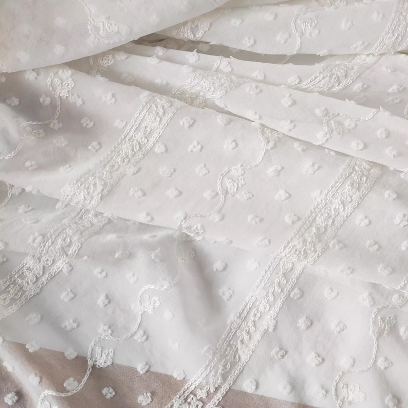 May include: White cotton fabric with a delicate embroidered pattern of small dots and floral designs. The fabric has a slightly textured surface.