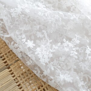 May include: White sheer fabric with delicate white floral embroidery. The fabric is soft and lightweight, perfect for creating garments or home decor.