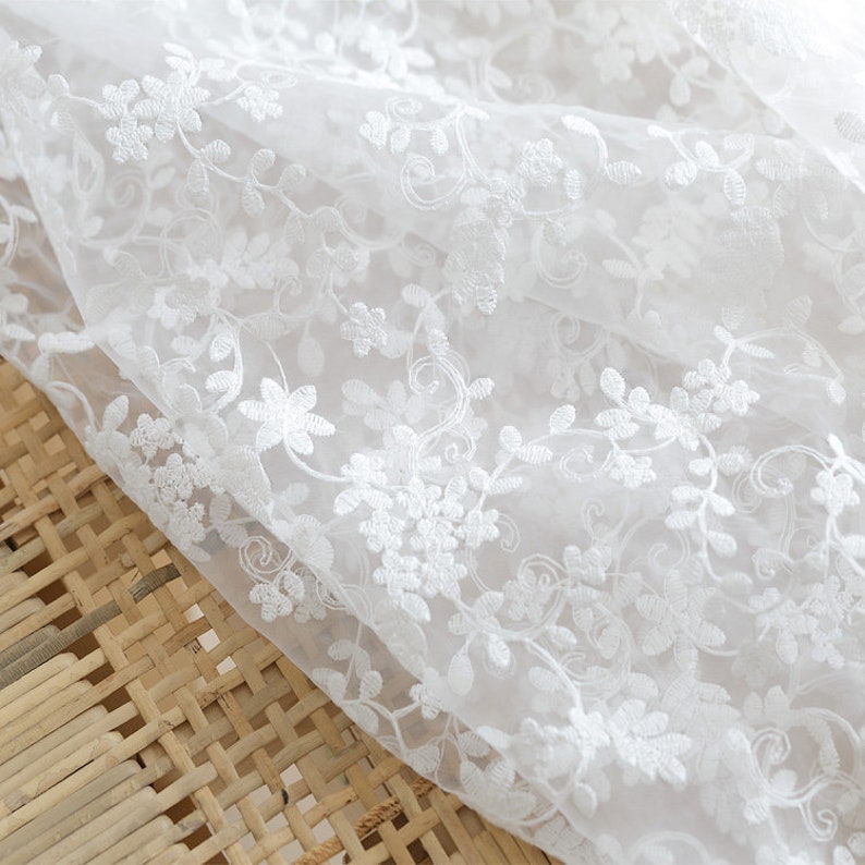 May include: White sheer fabric with delicate white floral embroidery. The fabric is soft and lightweight, perfect for creating garments or home decor.