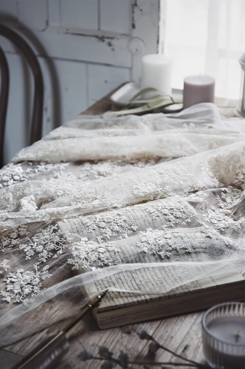Op de afbeelding: Witte kant met delicate bloemenborduursel, gedrapeerd over een boek en een houten tafel. De stof heeft een zachte, romantische uitstraling en zou perfect zijn voor een verscheidenheid aan knutselprojecten.